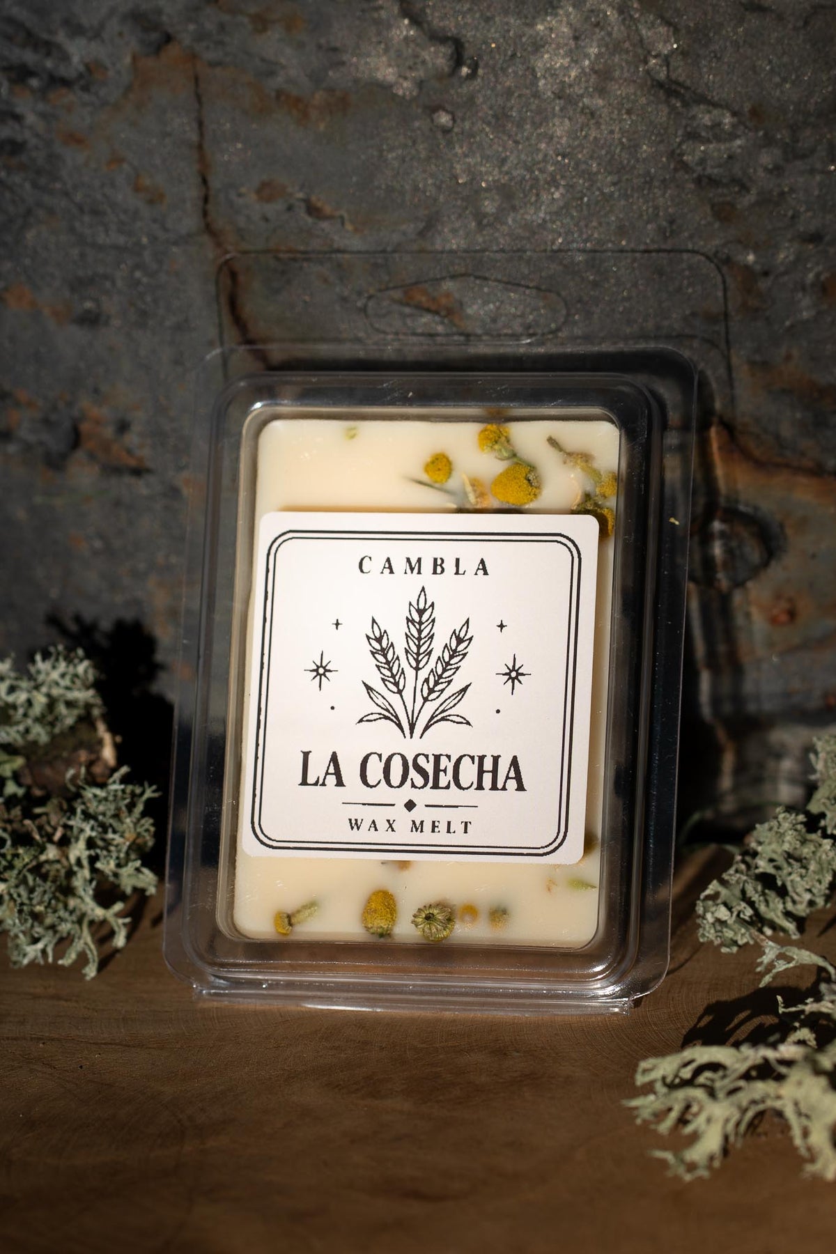 La Cosecha Wax Melt de Cambla con flores de manzanilla visibles en cera blanca, packaging transparente sobre madera con musgo