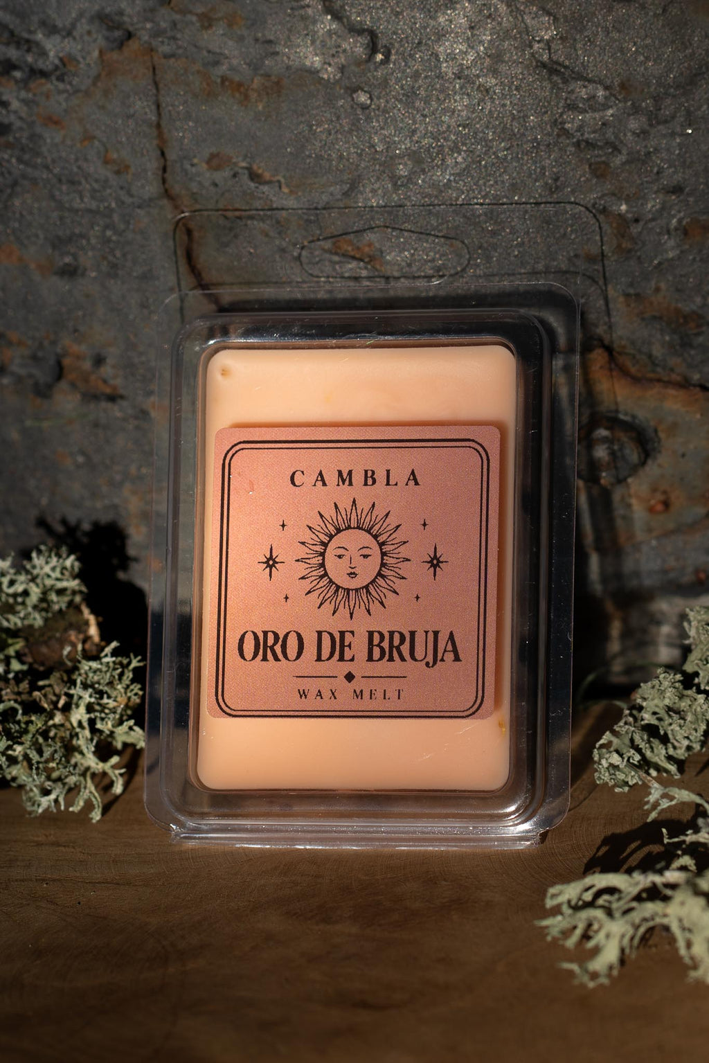 Oro de Bruja Wax Melt de Cambla en packaging transparente dorado rosado sobre madera con musgo