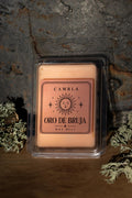 Oro de Bruja Wax Melt de Cambla en packaging transparente dorado rosado sobre madera con musgo