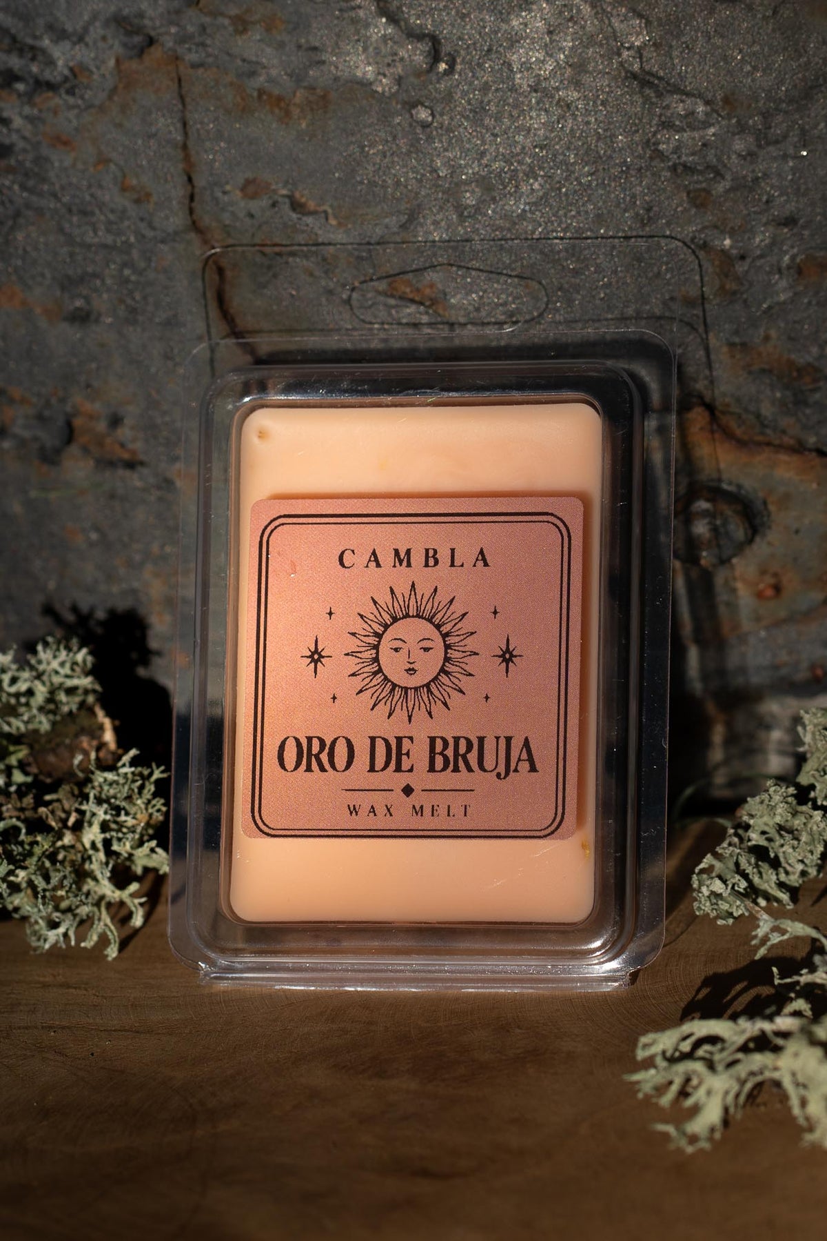 Oro de Bruja Wax Melt de Cambla en packaging transparente dorado rosado sobre madera con musgo